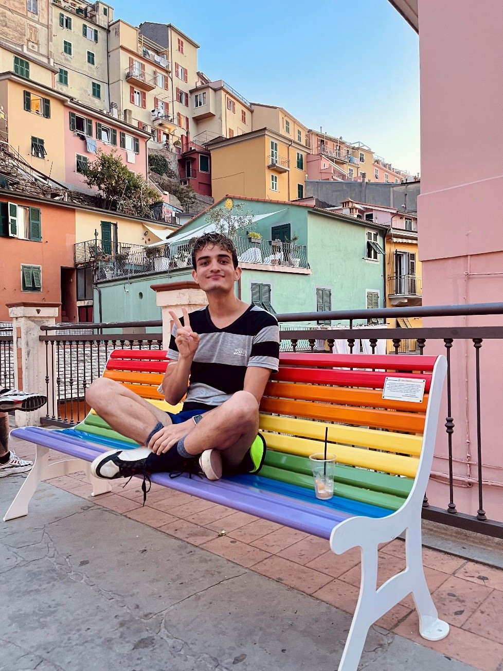 Gus is sitting on a rainbow bench. He is smiling and holding up a peace sign. 